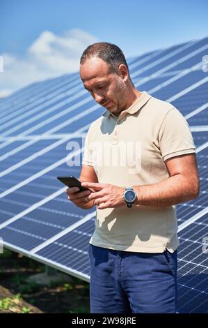 Geschäftiger Geschäftsmann, der mit allem Schritt halten will. Selbstbewusster Mann, der seine neue Investition überprüft. Junger Mann, der sein Handy auf dem Hintergrund eines Solarmoduls ansieht. Stockfoto