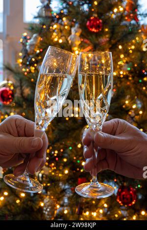 Silvester- oder weihnachtsessen, zwei Mannhände mit Gläsern Sekt Cava oder Prosecco in Kristallgläsern mit weihnachtsbaum- und Girlandenbeleuchtung Stockfoto