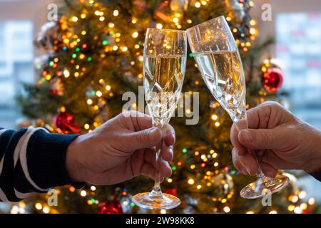Silvester- oder weihnachtsessen, zwei Mannhände mit Gläsern Sekt Cava oder Prosecco in Kristallgläsern mit weihnachtsbaum- und Girlandenbeleuchtung Stockfoto