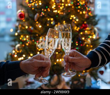 Silvester- oder weihnachtsessen, zwei Mannhände mit Gläsern Sekt Cava oder Prosecco in Kristallgläsern mit weihnachtsbaum- und Girlandenbeleuchtung Stockfoto