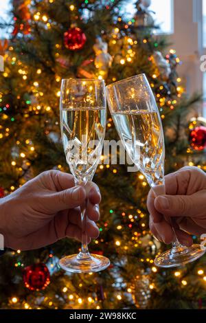 Silvester- oder weihnachtsessen, zwei Mannhände mit Gläsern Sekt Cava oder Prosecco in Kristallgläsern mit weihnachtsbaum- und Girlandenbeleuchtung Stockfoto