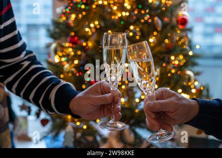 Silvester- oder weihnachtsessen, zwei Mannhände mit Gläsern Sekt Cava oder Prosecco in Kristallgläsern mit weihnachtsbaum- und Girlandenbeleuchtung Stockfoto