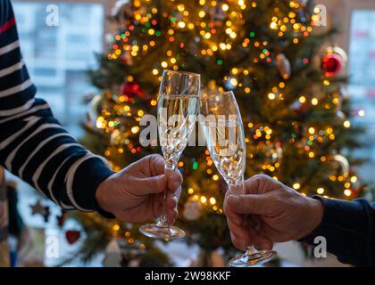 Silvester- oder weihnachtsessen, zwei Mannhände mit Gläsern Sekt Cava oder Prosecco in Kristallgläsern mit weihnachtsbaum- und Girlandenbeleuchtung Stockfoto