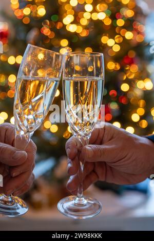 Silvester- oder weihnachtsessen, zwei Mannhände mit Gläsern Sekt Cava oder Prosecco in Kristallgläsern mit weihnachtsbaum- und Girlandenbeleuchtung Stockfoto