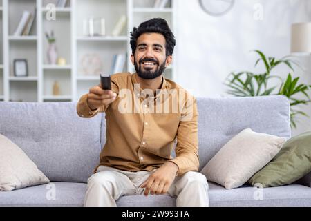 Ein lächelnder junger indischer Mann entspannt sich zu Hause, sitzt auf dem Sofa und schaltet den Fernseher mit der Fernbedienung ein. Stockfoto