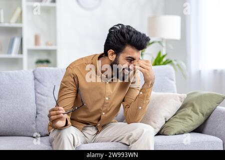 Ein junger indischer Mann sitzt zu Hause auf dem Sofa, hält eine Brille und lacht mit der Hand, die sein Gesicht bedeckt. Stockfoto