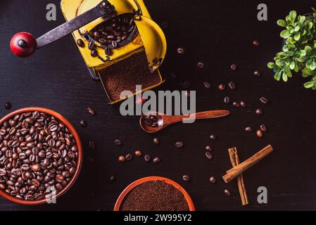 Kaffeemühle neben einigen Schüsseln mit gemahlenem Kaffee und Bohnen, Zimtstangen und einem Holzlöffel auf einem schwarzen Holztisch. Stockfoto