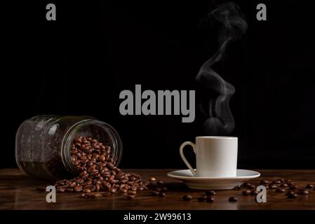 Tasse dampfenden Kaffee neben einem umgedrehten Glas mit Kaffeebohnen auf einem Holztisch mit mehr losen Bohnen, Vorderansicht. Stockfoto