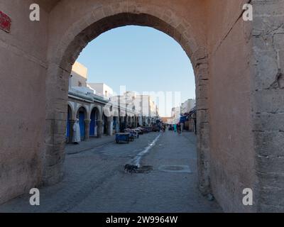 Die Hauptmedina-Straße mit einer Dachrinne im Zentrum, während eine Katze vorbeiläuft und ein Mann ein traditionelles Djellaba-Gewand in Essaouira, Marokko, trägt. Dezember 2023 Stockfoto