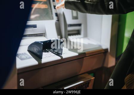 Stilvolle Leute, die an einem regnerischen Tag einen Geldautomaten benutzen, Geld abheben, während sie einen Regenschirm halten und ein Gespräch führen. Stockfoto