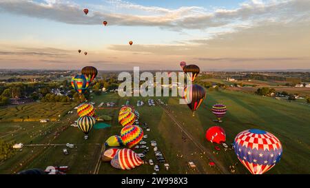 Aus der Vogelperspektive sehen Sie Heißluftballons in einem atemberaubenden Sonnenunterganghimmel, die während eines Festivals über ein üppiges grünes Gras- und Maisfeld schweben Stockfoto
