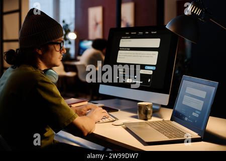 Ernsthafter junger KI-Designer, der sich den Computerbildschirm anschaut und die Tasten der Tastatur drückt, während er spät in der Nacht an einem neuen Projekt arbeitet Stockfoto