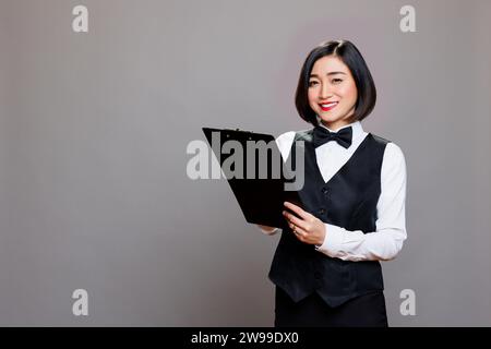 Attraktiv lächelnde asiatische Kellnerin, die auf eine Zwischenablage schreibt und die Kamera mit positivem Gesichtsausdruck betrachtet. Fröhliche junge Rezeptionistin in Uniform, die sich beim Posen im Studio-Porträt Notizen macht Stockfoto