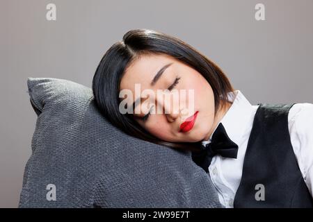 Erschöpfte, verschlafene asiatische Kellnerin, die Kopf auf Kissen ruhte und mit geschlossenen Augen im Studio posierte. Junge, attraktive, müde Rezeptionistin im Restaurant, die Uniform in der Nähe des Schlafens trug Stockfoto