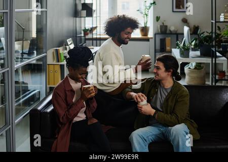 Drei junge kreative Designer trinken Kaffee und plaudern in der Pause, während sie auf einer schwarzen Ledercouch im Coworking Space sitzen Stockfoto