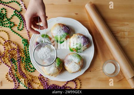 Über der Ansicht des weiblichen Handsiebens von Zuckerpulver auf appetitlichen Donuts auf einem Teller, der zwischen Papierrollen und Perlen steht Stockfoto