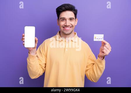 Foto-Porträt des gutaussehenden jungen Mannes halten weißen Bildschirm Kreditkarte gekleidet stilvolles gelbes Kleid isoliert auf lila Hintergrund Stockfoto