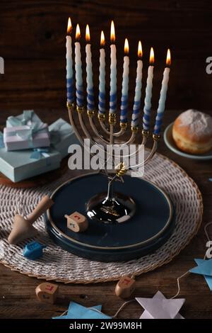 Hanukkah-Feier. Menora mit brennenden Kerzen, Dreidels, Geschenkboxen und Donut auf Holztisch Stockfoto