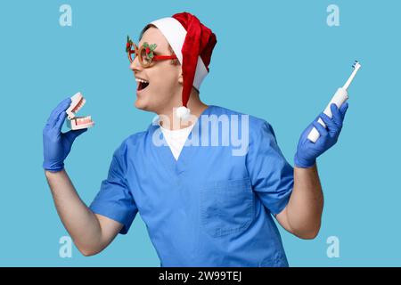 Porträt des jungen Zahnarztes im Weihnachtsmannmütze mit elektrischer Zahnbürste und Kiefermodell auf blauem Hintergrund Stockfoto