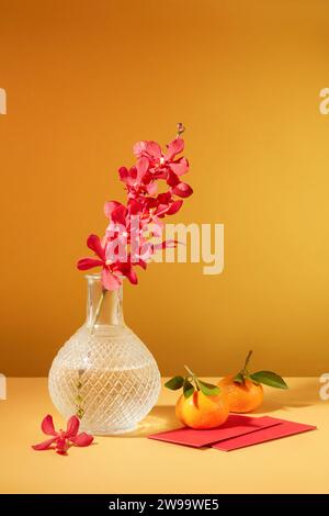 Eine Orchidee wird in eine transparente Glasvase gelegt, Mandarinen und Glücksgeldumschläge werden auf einem beigefarbenen Tisch mit gelbem Hintergrund ausgestellt. Stockfoto