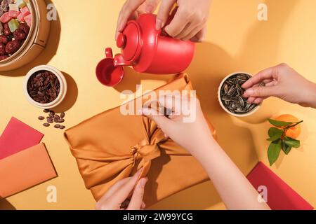 Eine Person wickelt ein Geschenk mit einem gelben Seidentuch ein, eine Person gießt Tee, die andere nimmt Sonnenblumenkerne auf. Tet-Teetisch mit Stockfoto