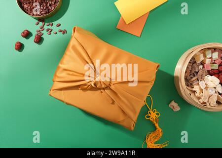 Blick von oben auf eine Geschenkbox in gelber Seide, ein Tablett mit Marmelade und Melonensamen und Glücksgeldumschläge auf grünem Hintergrund. Tet-Teetisch. Stockfoto