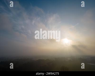 Das Bild fängt einen atemberaubenden Sonnenaufgang als zentralen Fokus ein, wobei die Sonnenstrahlen durch einen dramatischen Himmel mit Wolken durchdringen. Der Nebel, der über der ländlichen Landschaft dahinter liegt, wird beleuchtet und erzeugt ein diffuses Leuchten, das die Details des Geländes weicht. Die aufgehende Sonne bringt Wärme und verspricht einen neuen Tag in die kühlen, gedämpften Töne der frühen Morgenlandschaft. Die ätherische Qualität des Lichts und die Weite des Himmels erwecken ein Gefühl des Friedens und der Pracht der Natur. Sonnenaufgang Majestät über einer nebeligen ländlichen Landschaft. Hochwertige Fotos Stockfoto