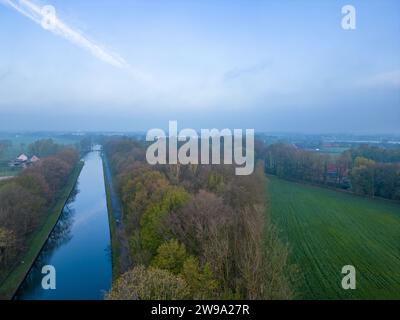 Dieses ruhige Bild fängt einen nebeligen Morgen über einem ruhigen Kanal ein, flankiert von herbstlichen Baumreihen und üppigen grünen Feldern. Das sanfte Licht der Dämmerung beleuchtet die Szene sanft und erzeugt einen subtilen Verlauf am Himmel, während ein Hauch von Nebel eine geheimnisvolle Schicht hinzufügt. Der Kanal führt den Blick zum Horizont und lädt zum Betrachten der Distanz und des ruhigen ländlichen Lebens darüber hinaus ein. Nebeliger Morgen über Kanal und Felder. Hochwertige Fotos Stockfoto