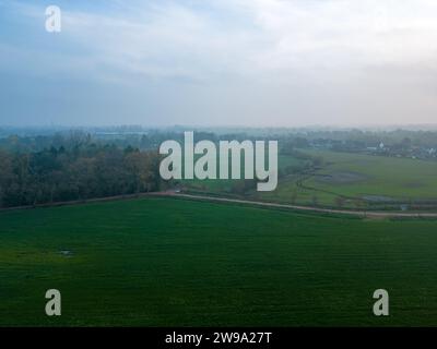 Das Bild fängt das heitere Wesen eines nebeligen morgens über einem weitläufigen Ackerland ein. Das sanfte Licht der Morgendämmerung durchströmt den Dunst und wirft ein sanftes Leuchten über die grünen Felder. Der ferne Horizont ist kaum zu sehen, umhüllt vom Morgennebel, was der Landschaft eine ätherische Qualität verleiht. Die geschwungenen Linien des Geländes und die subtilen Grüntöne schaffen eine friedliche, pastorale Szene, die ein Gefühl der Ruhe und das langsame Erwachen der Landschaft hervorruft. Misty Dawn Über Sanftes Rollendes Ackerland. Hochwertige Fotos Stockfoto