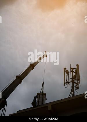 Industriekrane zum Betrieb und Anheben der elektrischen Box gegen Wolken und Himmel mit Telekommunikationsturm Stockfoto