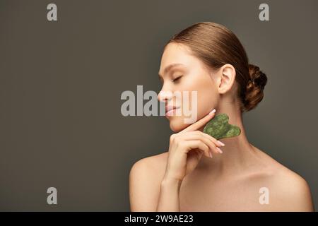 Junge Frau, die Gesichtsmassage mit Jade-Gua-Sha-Stein auf grauem Hintergrund macht, Gesichtspflegekonzept Stockfoto