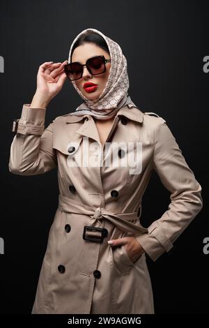 Stilvolles weibliches Modell im Trenchcoat und Kopftuch, das vor der Kamera posiert. Porträt einer modischen Frau mit Kopfstück, Sonnenbrille, während sie vor schwarzem Hintergrund steht. Modekonzept. Stockfoto