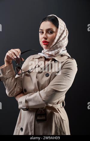 Wunderschöne, rot lippige Frau im trendigen Outfit, die nachdenklich in die Kamera schaut. Porträt einer schönen kaukasischen Dame im Trenchcoat, die über dunklem Hintergrund posiert. Konzept der Modellierung, Mode, Stil. Stockfoto