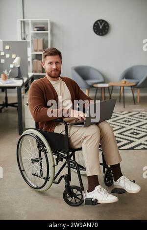 Vertikales Vollbildnis eines bärtigen Mannes mit Behinderung mit Laptop im Rollstuhl und Blick auf die Kamera Stockfoto