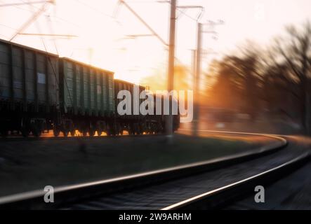 Güterzug, der bei Sonnenuntergang vorbeifährt. Zug mit Fracht unter den Sonnenstrahlen der untergehenden Sonne. Bewegungsunschärfe. Weichzeichner. Stockfoto