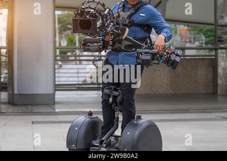 Video-, Filmemachen- und Kreativkonzept - Nahaufnahme einer modernen Filmkamera auf 3-Achsen-Gimbal-Stabilisator in männlichen Händen Gehen Sie auf einem Segway. Stockfoto