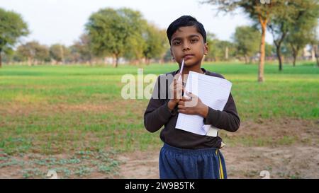 Lächelnder Student Junge mit Heft und Bleistift in der Hand. Porträt des denkenden asiatischen Dorfjungen auf grünem Farmland. Stockfoto