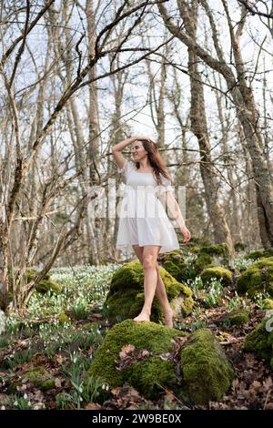 Schneeglöckchen galanthus-Frau. Sie steht in einem weißen Kleid auf einer Wiese mit Schneeglöckchen in einem Frühlingswald Stockfoto