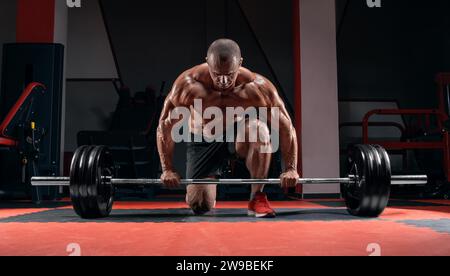 Der Athlet steht auf seinem Knie und in der Nähe der Bar im Fitnessstudio und bereitet sich auf einen Deadlift vor. Gemischte Medien Stockfoto