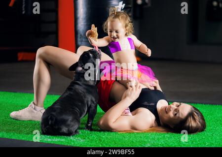 Die charmante Sportmutter spielt im Fitnessstudio mit ihrer kleinen Tochter und einer französischen Bulldogge. Gemischte Medien Stockfoto