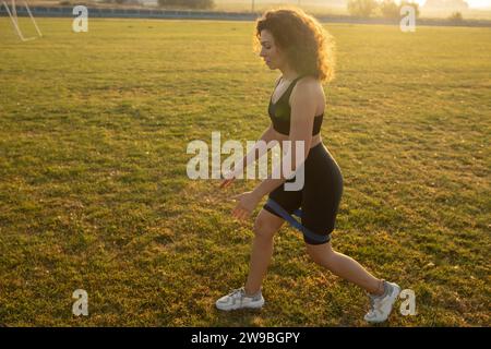 Junge lockige Sportmädchen in Sportbekleidung führt Ausfallschritte mit Widerstandsband draußen auf dem Gras bei Sonnenuntergang durch Stockfoto