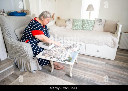 Eine ältere Frau sammelt Puzzle, während sie in einem Stuhl in ihrem hellen Zimmer zu Hause sitzt. Stockfoto