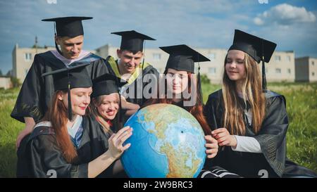 Absolventen in schwarzen Gewändern untersuchen einen geografischen Globus, der auf dem Gras sitzt. Stockfoto
