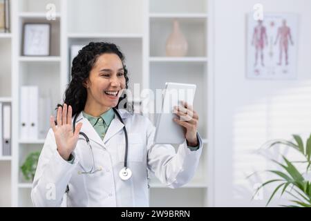 Junge hispanische Ärztin, die im Klinikbüro steht und mit dem Patienten über Videoanrufe spricht, sich online berät, begrüßt und mit der Tablet-Kamera winkt. Stockfoto