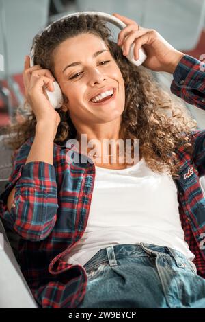 Glückliche junge Frau mit lockigen Haaren in kariertem Hemd, die auf der Couch sitzt und Kopfhörer anpasst, während sie während der Freizeit zu Hause Lieder genießt Stockfoto