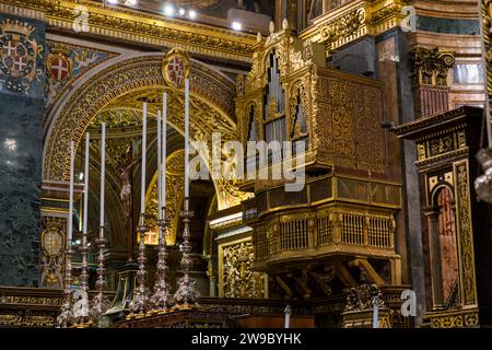 Eine vergoldete Orgelgalerie mit maltesischen Kreuzmedaillons erhebt sich über einem Altarbereich, der mit Kerzenständern und einem Kruzifix geschmückt ist, in der St. Johannes-Ko-Kathedrale in Valletta, Malta. Der Orden von St. Johann ließ die Kathedrale zwischen 1572 und 1577 erbauen; ihr reich vergoldetes Interieur stammt größtenteils aus den 1660er Jahren und später durch die Arbeit von Mattia Preti. Stockfoto