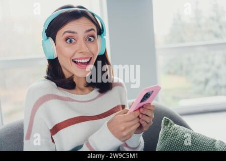 Porträt der erstaunten fröhlichen Dame Sessel Verwenden Sie Smartphone Musik Kopfhörer Zeitvertreib Chill Flat Indoor Stockfoto