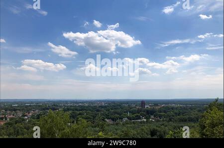 Dieses Bild bietet einen weiten Blick aus einem hohen Blickwinkel und einen Blick auf den weiten Horizont. Die grüne Landschaft erstreckt sich weit und breit, mit Bäumen und Gebäuden, die sich in die Ferne verschmelzen. Über dem Himmel ist eine Leinwand aus sanftem Blau, verziert mit flauschigen Wolken, die sanfte Schatten über das Gelände werfen und die Ruhe eines klaren Tages verkörpern. Stockfoto