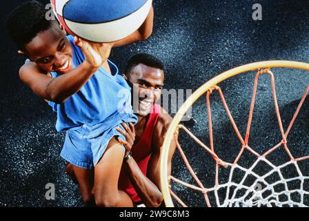 Ein afroamerikanischer Vater hebt seinen Sohn an, wie man einen Korb bei einem Basketballspiel gewinnt Stockfoto