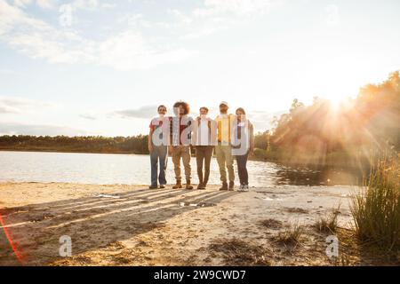 Das Bild zeigt eine entspannte und zufriedene Gruppe von Freunden, die zusammen an einem See stehen und hinter ihnen die Sonne untergeht. Die Gruppe ist vielfältig, darunter ein junger kaukasischer Junge, ein Mann aus dem Nahen Osten, eine hispanische Frau und ein älterer kaukasischer Mann. Das sanfte Abendlicht wäscht über sie, schafft eine ruhige Atmosphäre und wirft lange Schatten auf den Boden. Der offene Raum und die natürliche Umgebung wecken ein Gefühl von Frieden und Gesellschaft. Lakeside Unity: Die Golden Hour-Versammlung einer Gruppe. Hochwertige Fotos Stockfoto